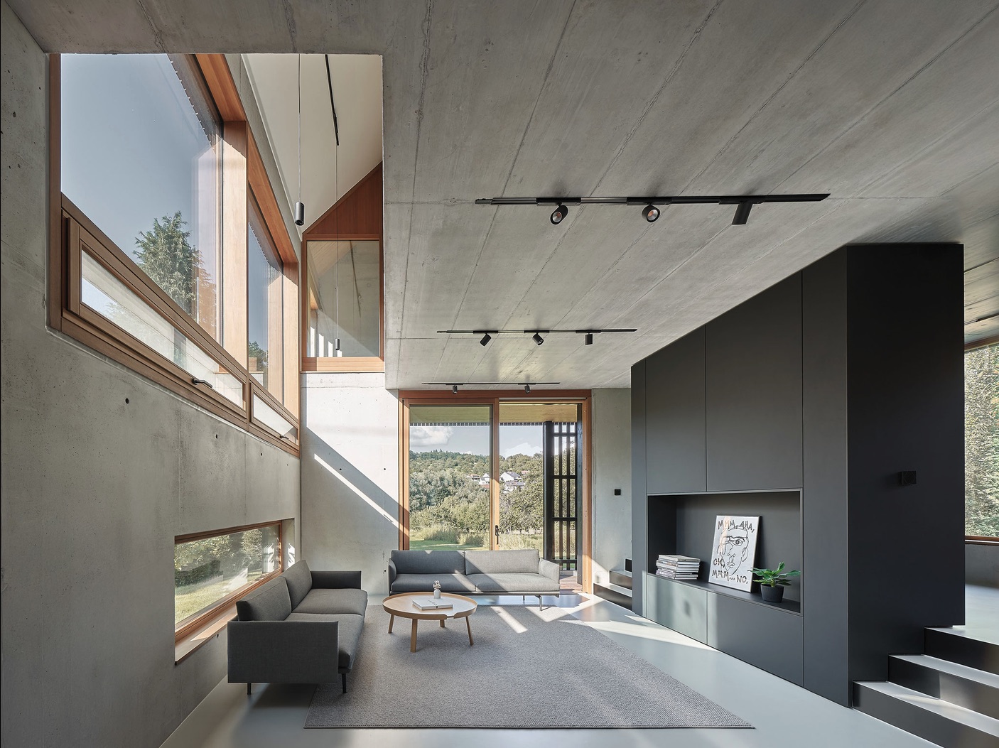 Haus RO – Wohnbereich mit grauen Sofas, raumhohen Holzfenstern und dunkler Möbelwand